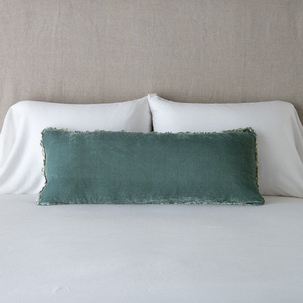 A rectangular green throw pillow with a plush texture, featuring a ruffled edge and tie closure, placed on a bed with white bedding.