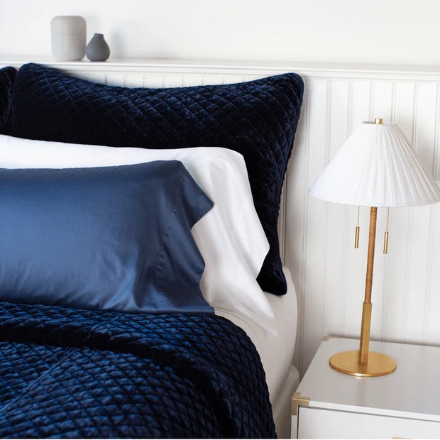 Navy blue quilted silk velvet sham with satin piping on a bed, with a bedside lamp and white walls.