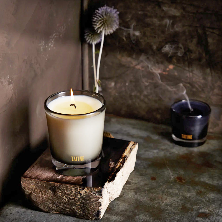An 8 oz natural wax candle in a smoke grey mouth-blown glass jar, with a cotton wick, sitting on a wooden surface, with a dark background.