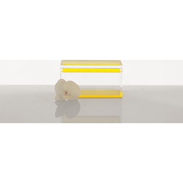 A clear acrylic Alexandra Von Furstenberg Treasure Box with a yellow detail on the lid, displayed alongside a white orchid flower.