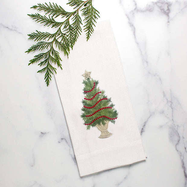 A white linen towel with a green embroidered Christmas tree design, displayed on a marble surface with a sprig of evergreen leaves to the side.