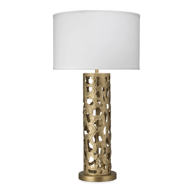 A gold leaf table lamp with a white linen drum shade, featuring intricate cut-outs in the base, on a white background.