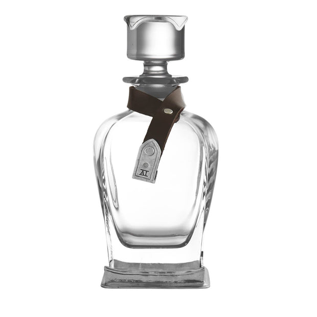 A clear glass liquor decanter with a pewter ornament and a brown leather strap around the neck, standing on a square base.