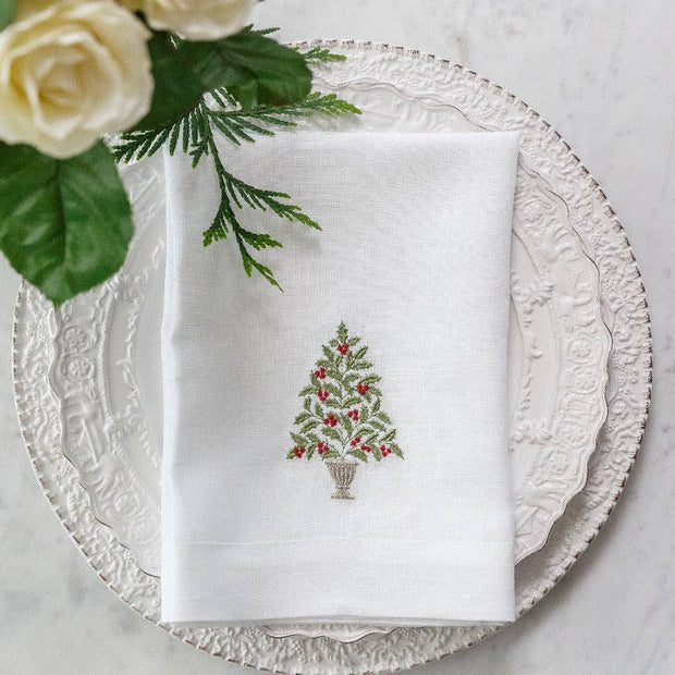 A white linen napkin with a green and red embroidered holly tree design, displayed on a white plate with a green and white flower arrangement.