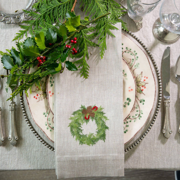 A linen towel with a embroidered wreath design, displayed on a table setting with cutlery and greenery in the background.