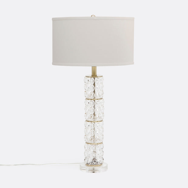 A tall and slender Beroe Table Lamp with clear glass panels and brass bands, set on an acrylic base, with a white lampshade.
