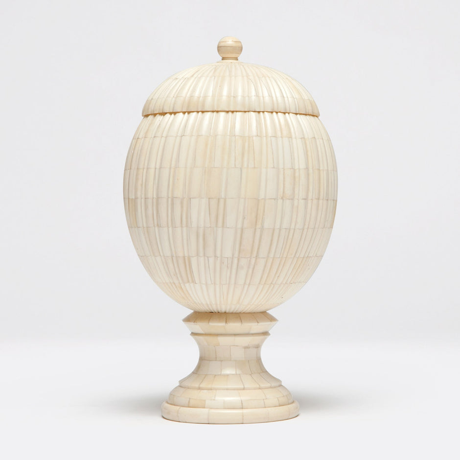 A bone-made decorative object resembling a pinecone with vertical ribs, featuring a curved base and lid, placed on a pedestal.