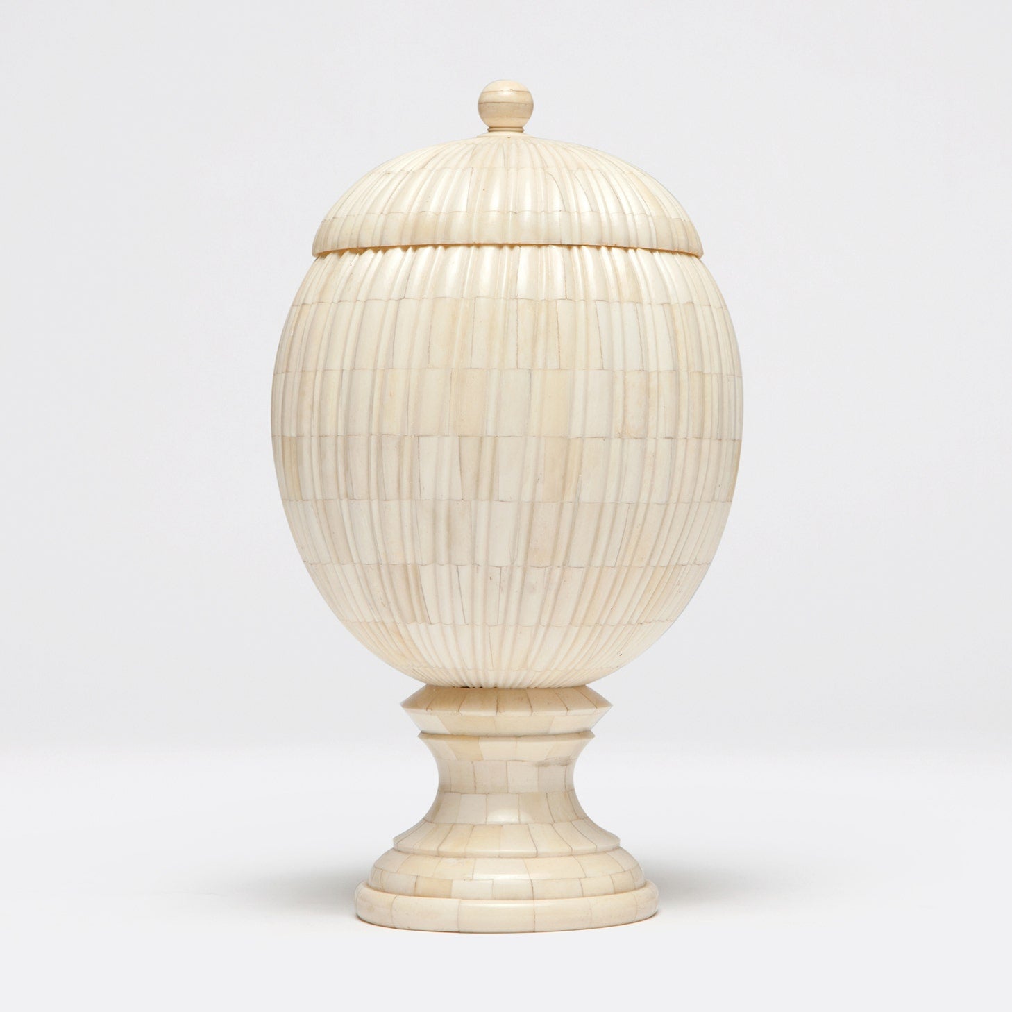 A bone-made decorative object resembling a pinecone with vertical ribs, featuring a curved base and lid, placed on a pedestal.