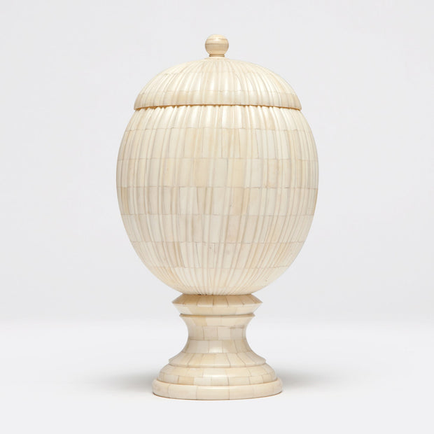 A bone-made decorative object resembling a pinecone with vertical ribs, featuring a curved base and lid, placed on a pedestal.