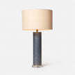 A cylindrical Tavis table lamp with a faux linen covering, brass discs on the top and bottom, and a circular crystal base, accompanied by a beige lampshade.