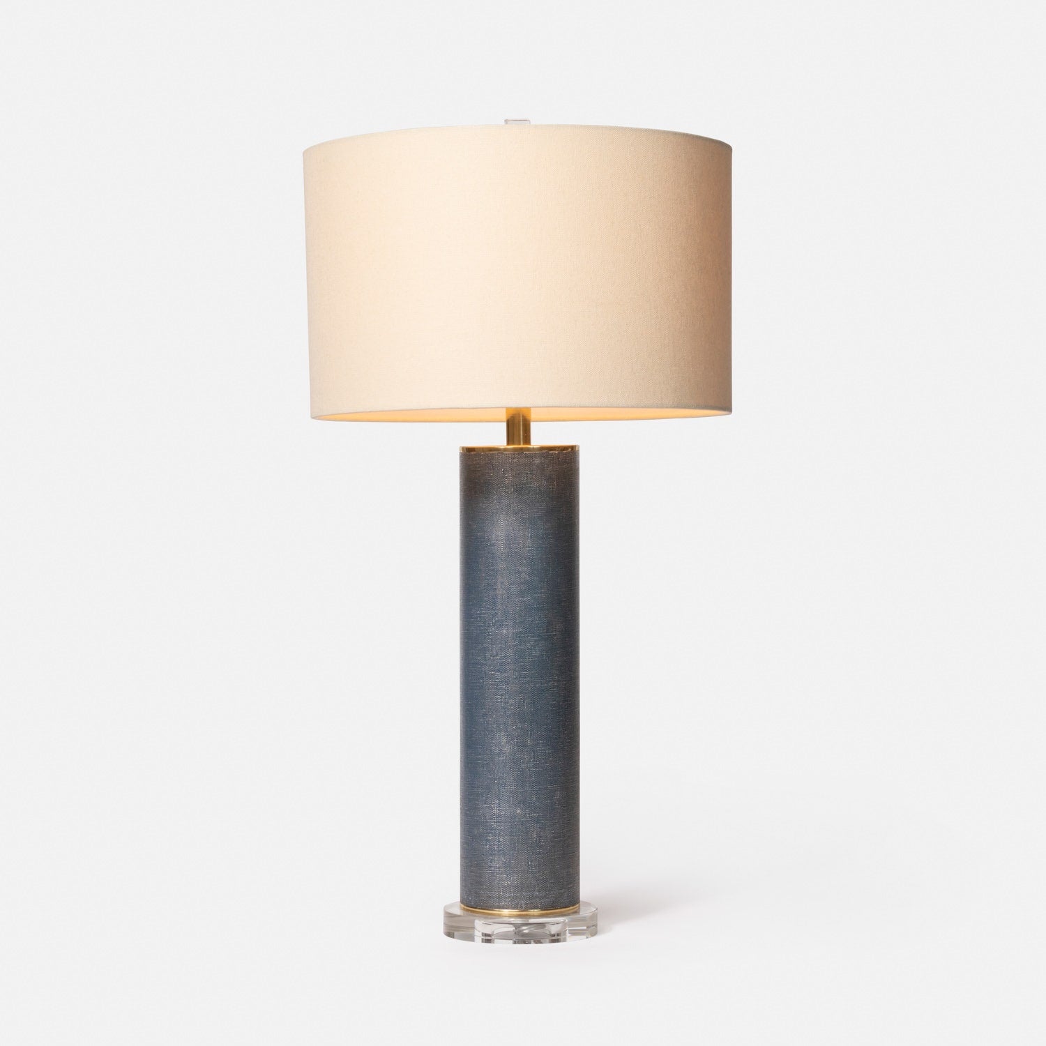A cylindrical Tavis table lamp with a faux linen covering, brass discs on the top and bottom, and a circular crystal base, accompanied by a beige lampshade.