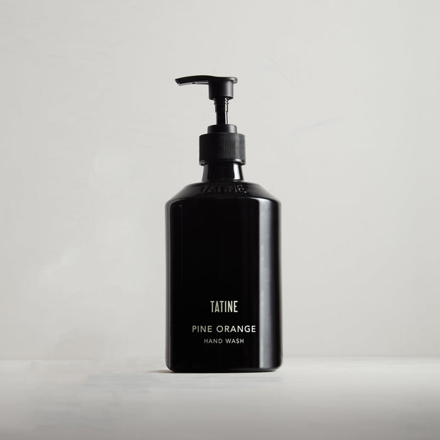 A black glass bottle with a pump dispenser labeled 'Tatine Pine Orange Hand Wash' on a light background.
