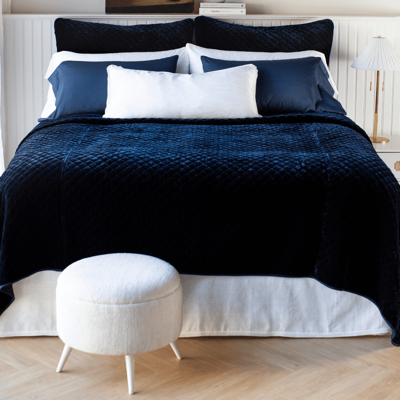 A luxurious dark blue silk velvet coverlet with a quilted pattern and satin edge, displayed on a bed with white and light blue bedding.