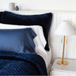 Navy blue quilted silk velvet sham with satin piping on a bed, with a bedside lamp and white walls.
