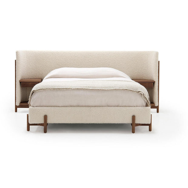 A king-sized Sullivan bed by Verellen with a natural walnut base and off-white upholstery, featuring an expansive headboard with integrated shelves and a side table.