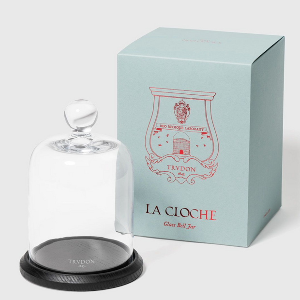 A clear glass cloche with a wooden base, featuring the Trudon brand logo, displayed next to its blue packaging box with the text 'LA CLOCHE' and the brand name.