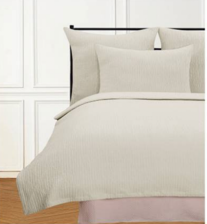 A white coverlet with a textured herringbone pattern on a bed, accompanied by two pillows, in a room with wooden flooring and a window in the background.