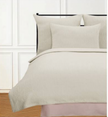 A white coverlet with a textured herringbone pattern on a bed, accompanied by two pillows, in a room with wooden flooring and a window in the background.