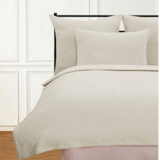 A white coverlet with a textured herringbone pattern on a bed, accompanied by two pillows, in a room with wooden flooring and a window in the background.